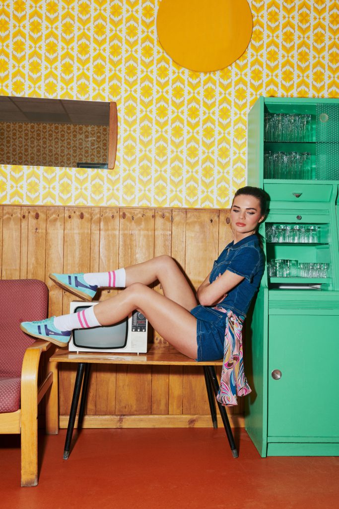 A woman sits on a table dressed in a denim dress and wearing Tornado trainers - retro aesthetic